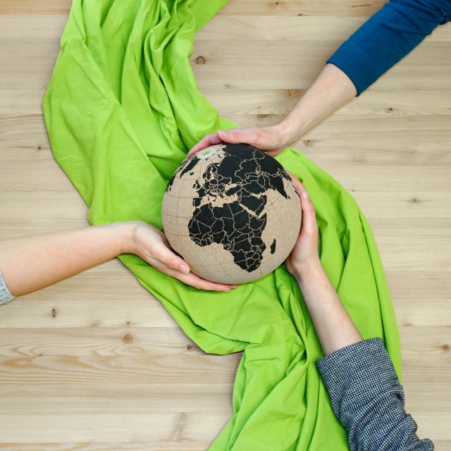 Drei Hände halten eine Erdkugel, die auf einem grünen Tuch am Boden oder einem Tisch laus Holz liegt.