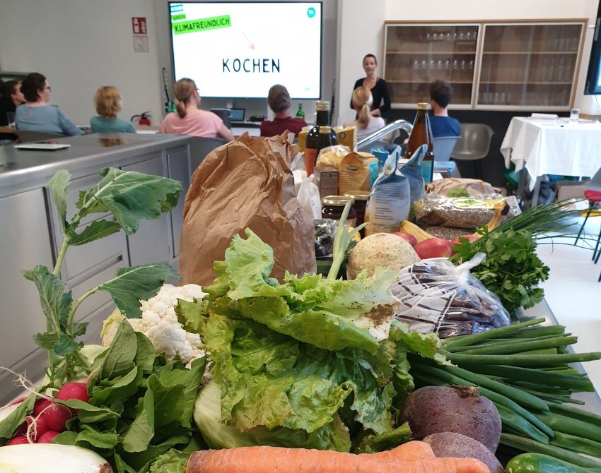 Salat, Frühlingszwiebel, Käferbohnen und weiteres Gemüse liegen auf einer Küchenzeile. Im Hintergrund eine Person im Stehen auf die sitzenden Personen schauen