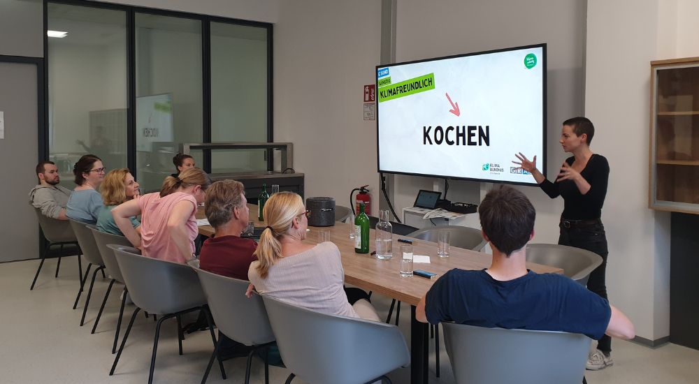 8 Menschen sitzen vor einem Tisch und schauen auf eine Person neben einem Screen, die etwas erklärt