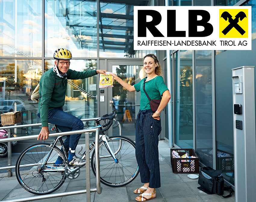 Älterer Mann auf Fahrrad nimmt Frühstückssackerl von junger Frau entgegen. Logo der Raiffeisen Landesbank Tirol AG
